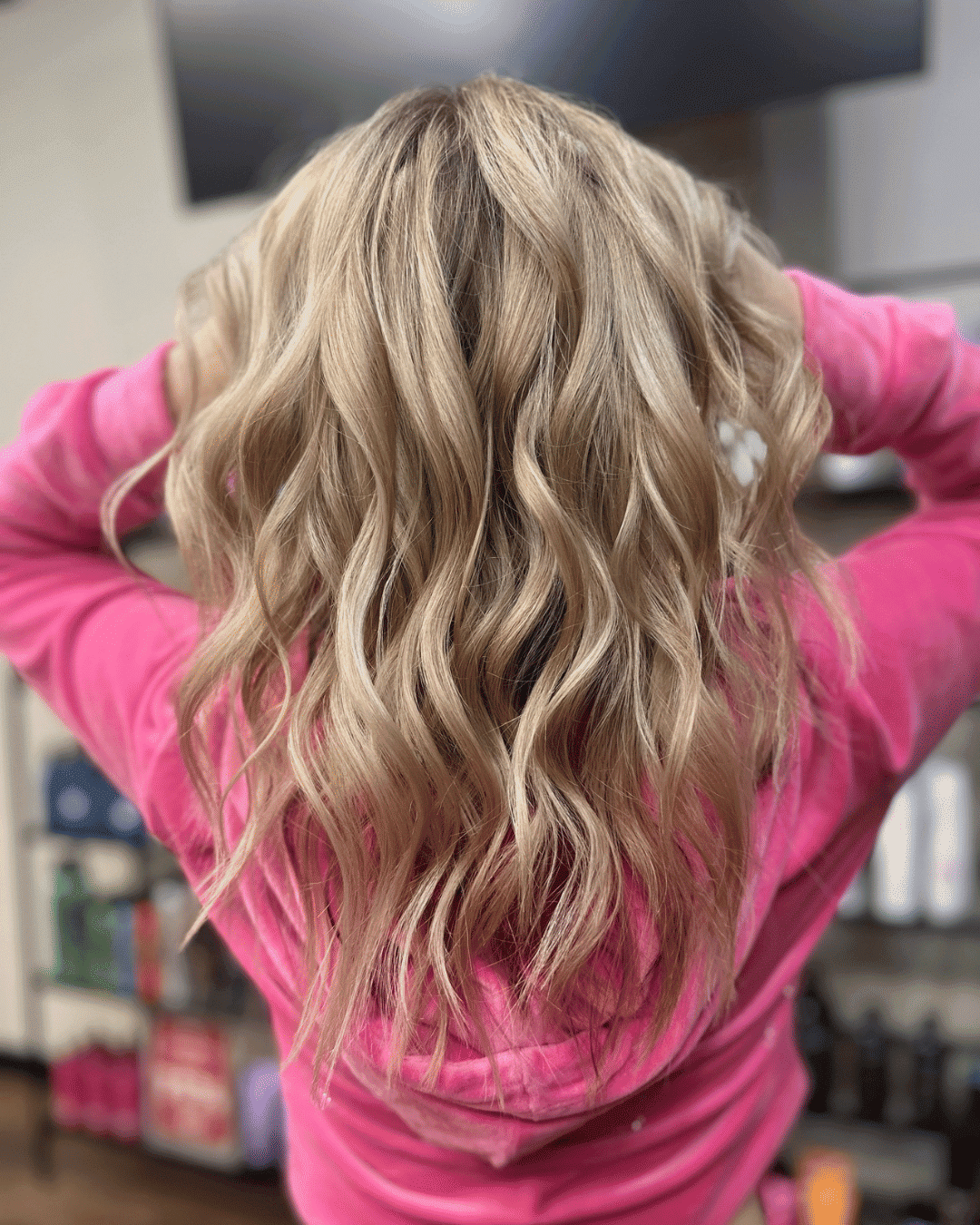 Back view of a person with wavy blonde hair, wearing a pink hoodie, in a salon setting.