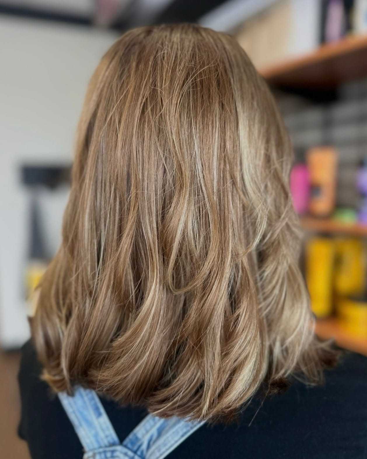 Brown hair with subtle highlights, styled in soft waves, framed by blurred background shelves.