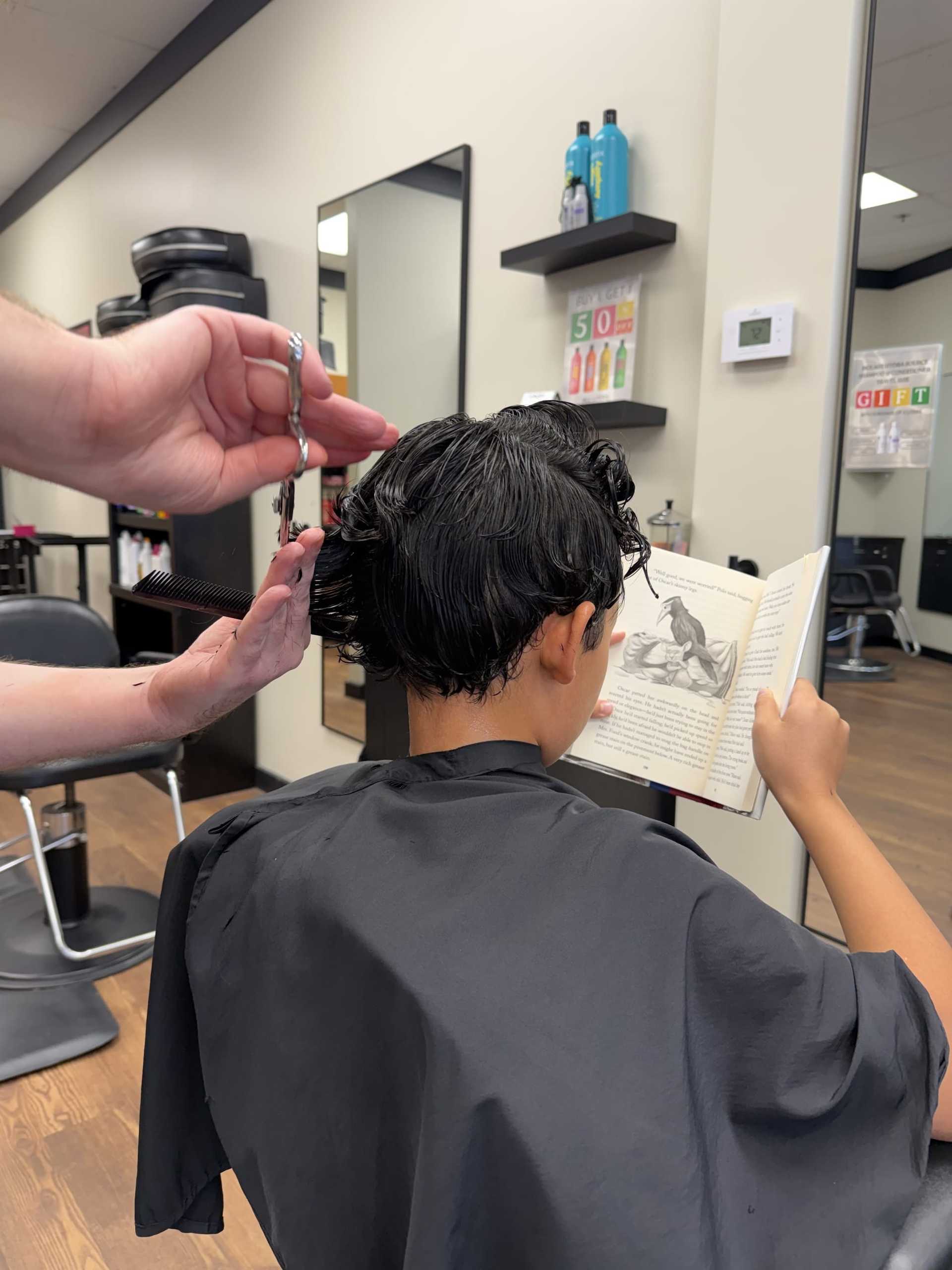 Young person reading book while getting a haircut in a salon.