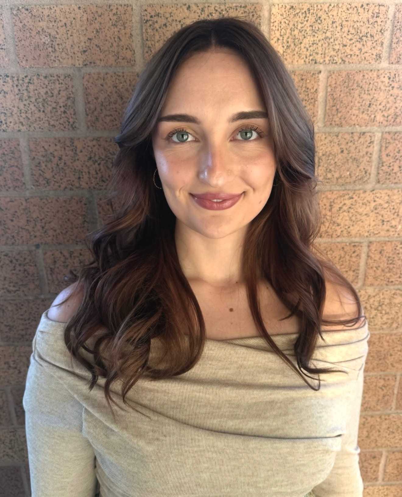 A woman with long brown hair and an off-shoulder top stands against a brick wall, smiling.