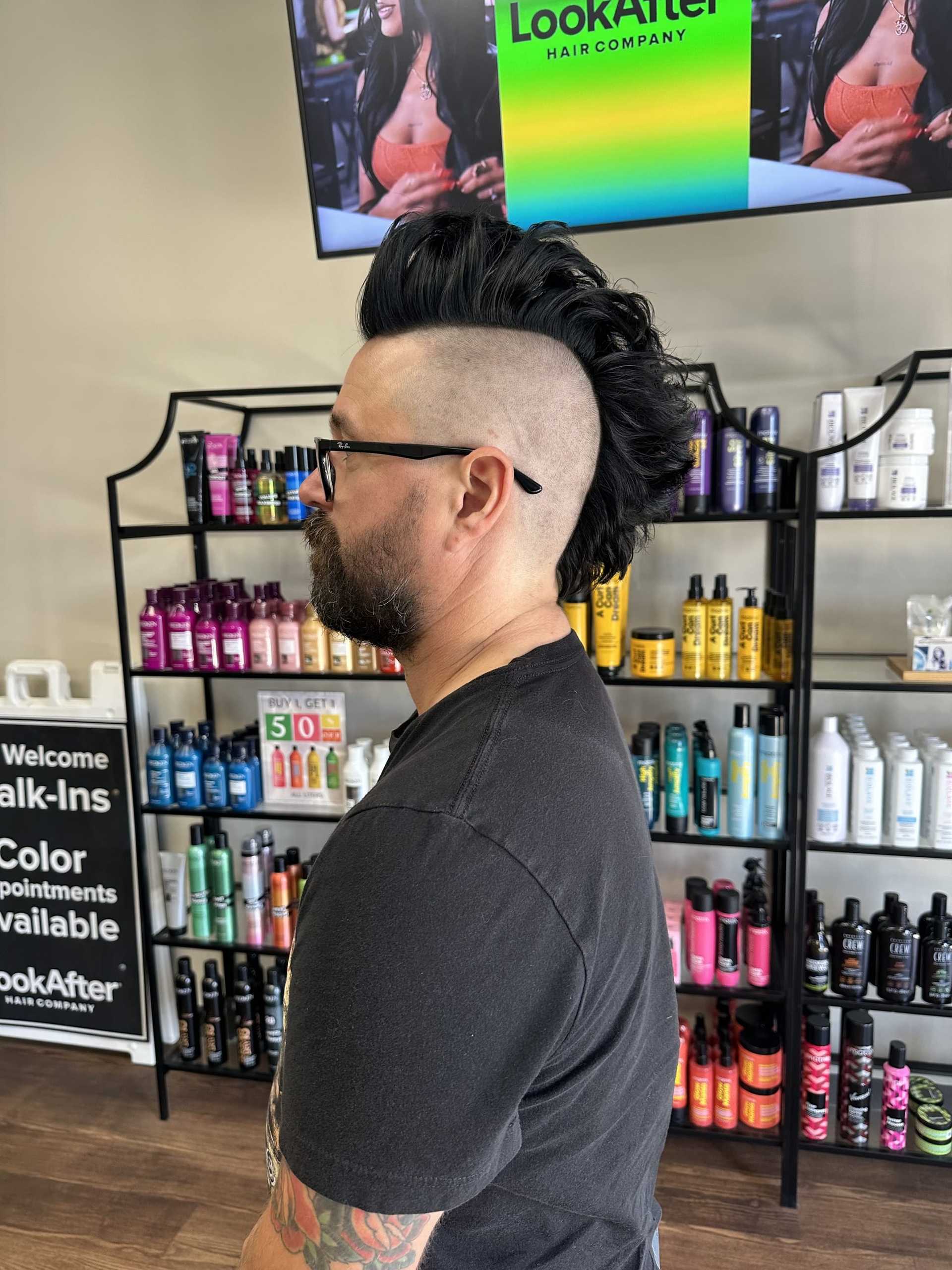 Man with mohawk hairstyle in salon, hair products displayed on shelves in background.
