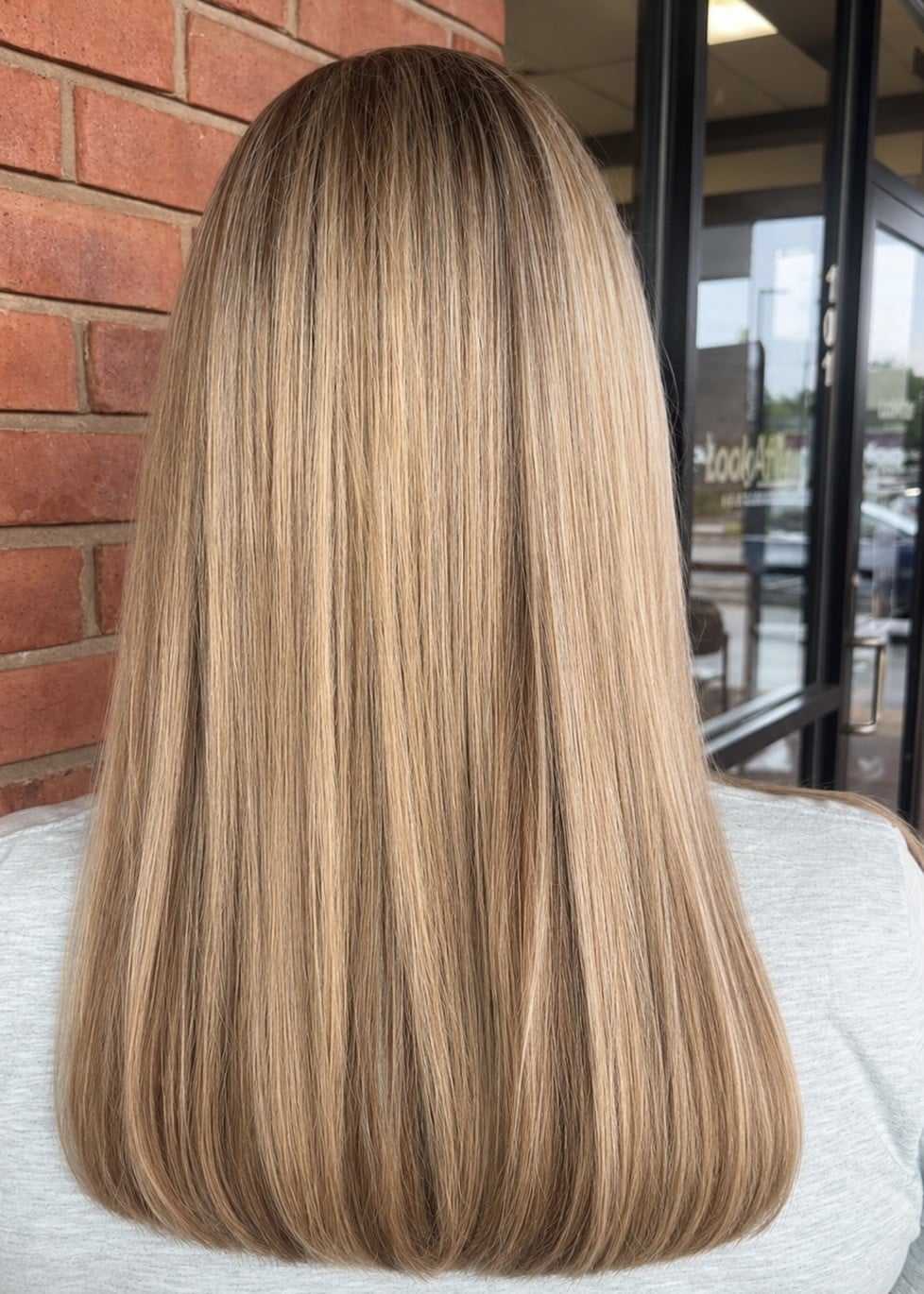 Straight, silky blonde hair with highlights, seen from the back, against a brick wall background.