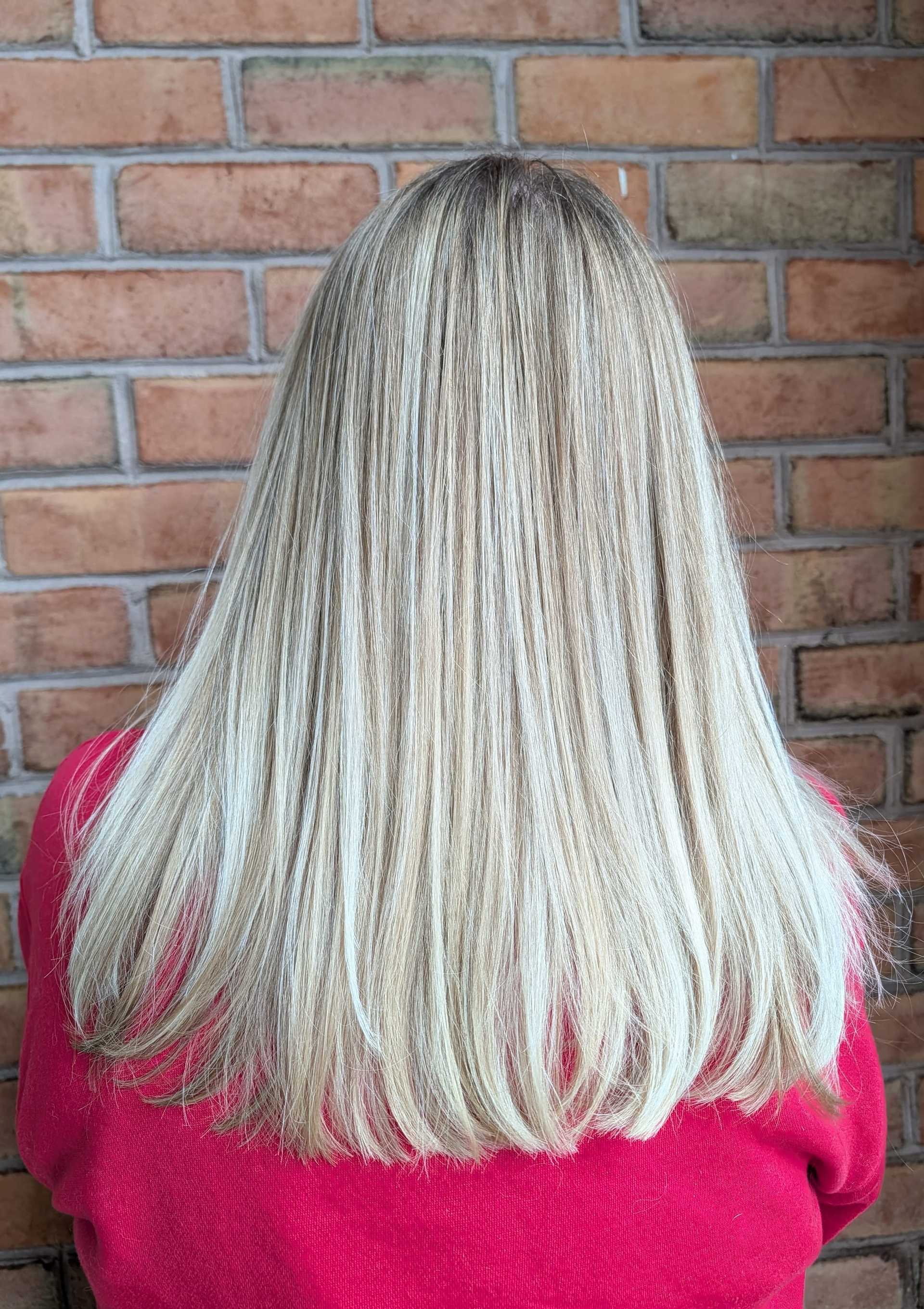 Back view of a person with straight, shoulder-length blond hair against a brick wall.