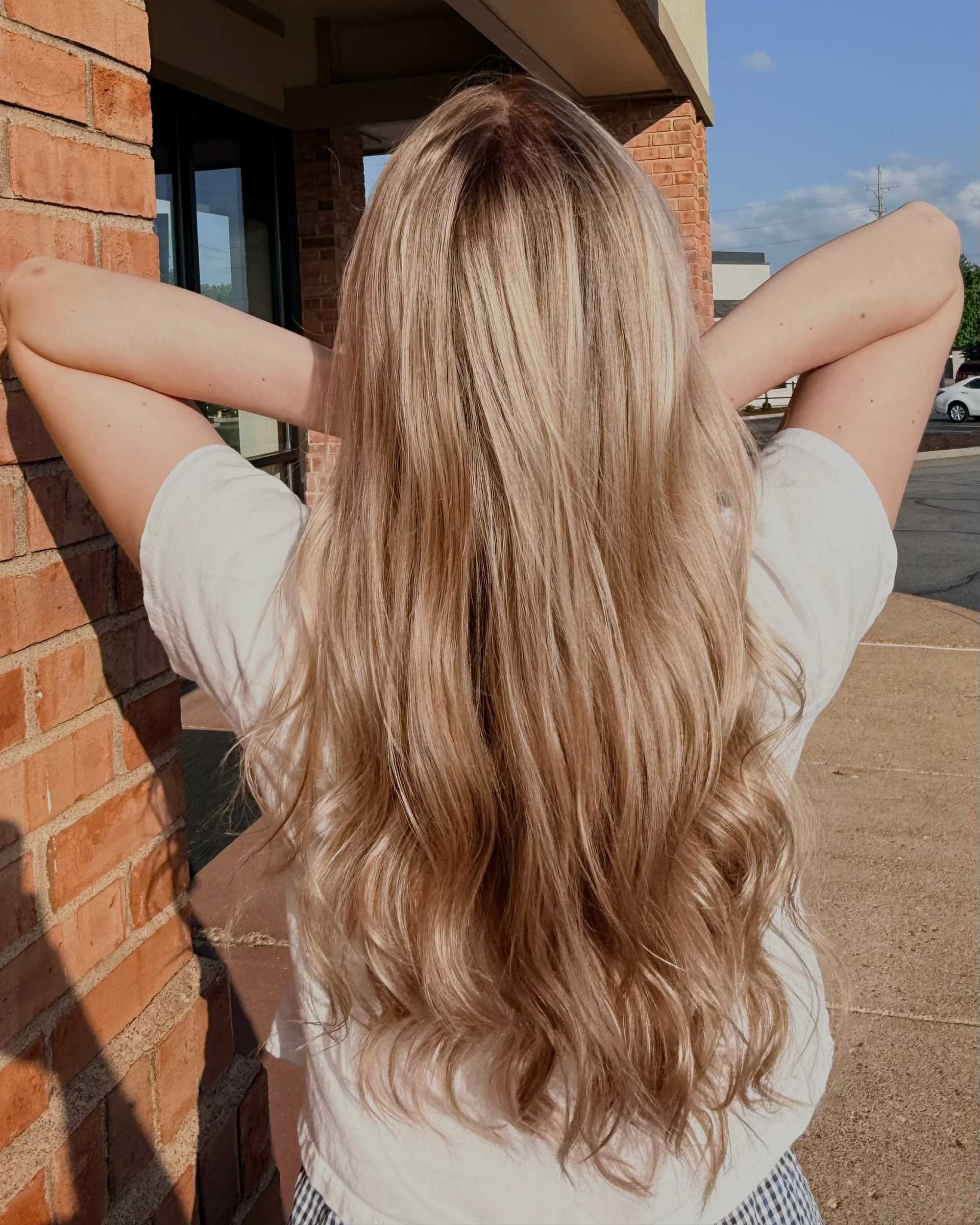 Long, wavy blonde hair flowing down the back of a person standing outside near a brick wall.