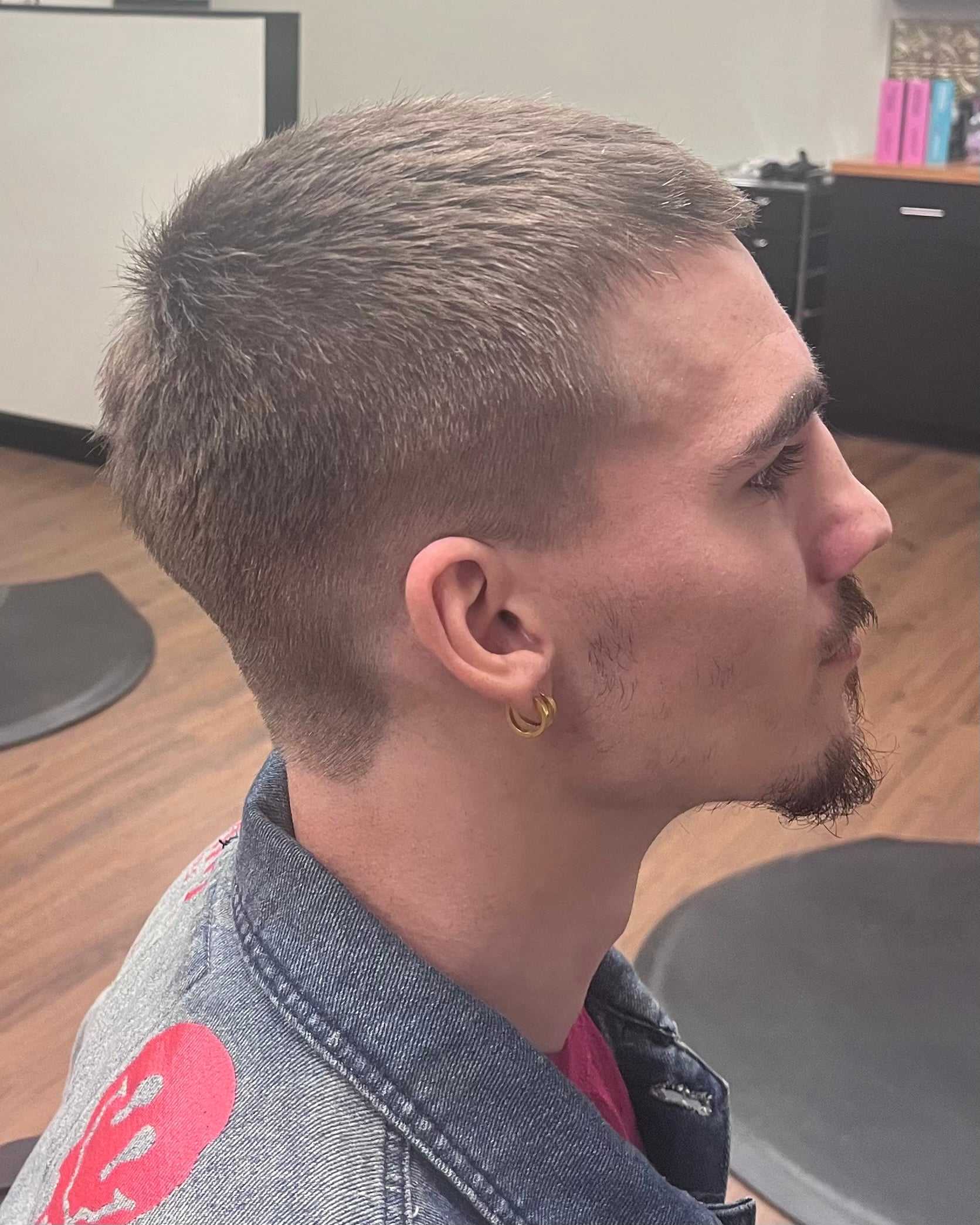 Man with short haircut and earrings, wearing a denim jacket in a salon setting.