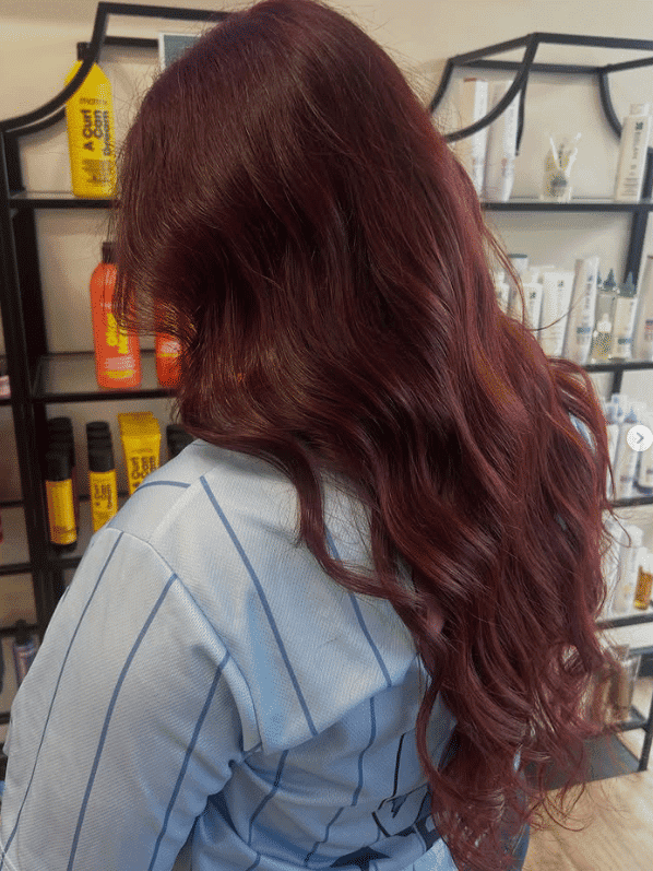 Woman with long, wavy red hair in a salon with hair products on shelves.