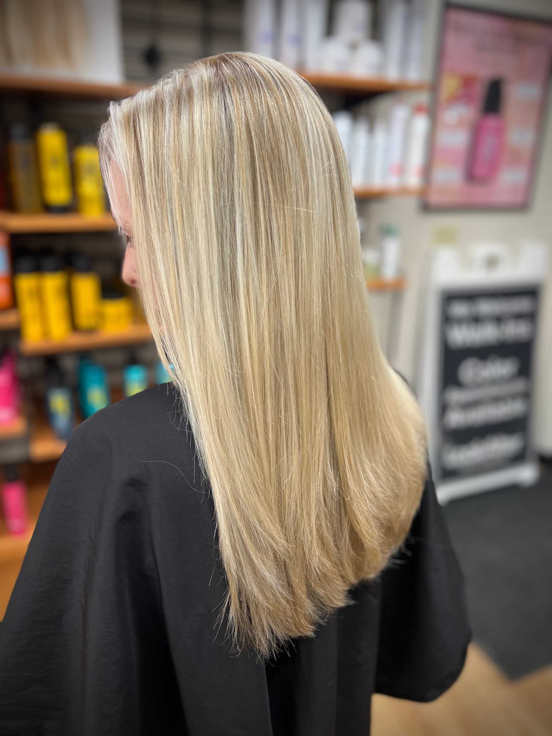 Blonde hair with highlights on a person in a salon, shelves of hair products behind.