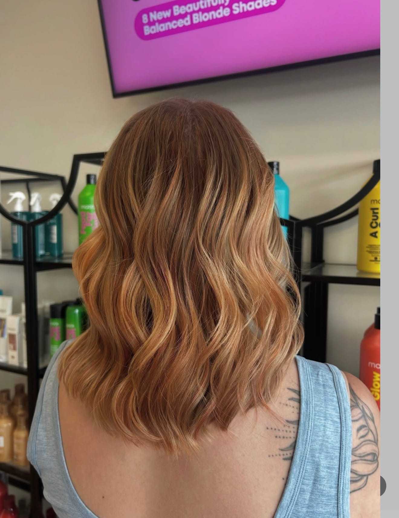 Red-haired person with wavy, blonde-highlighted hair in a salon, bottles in the background.