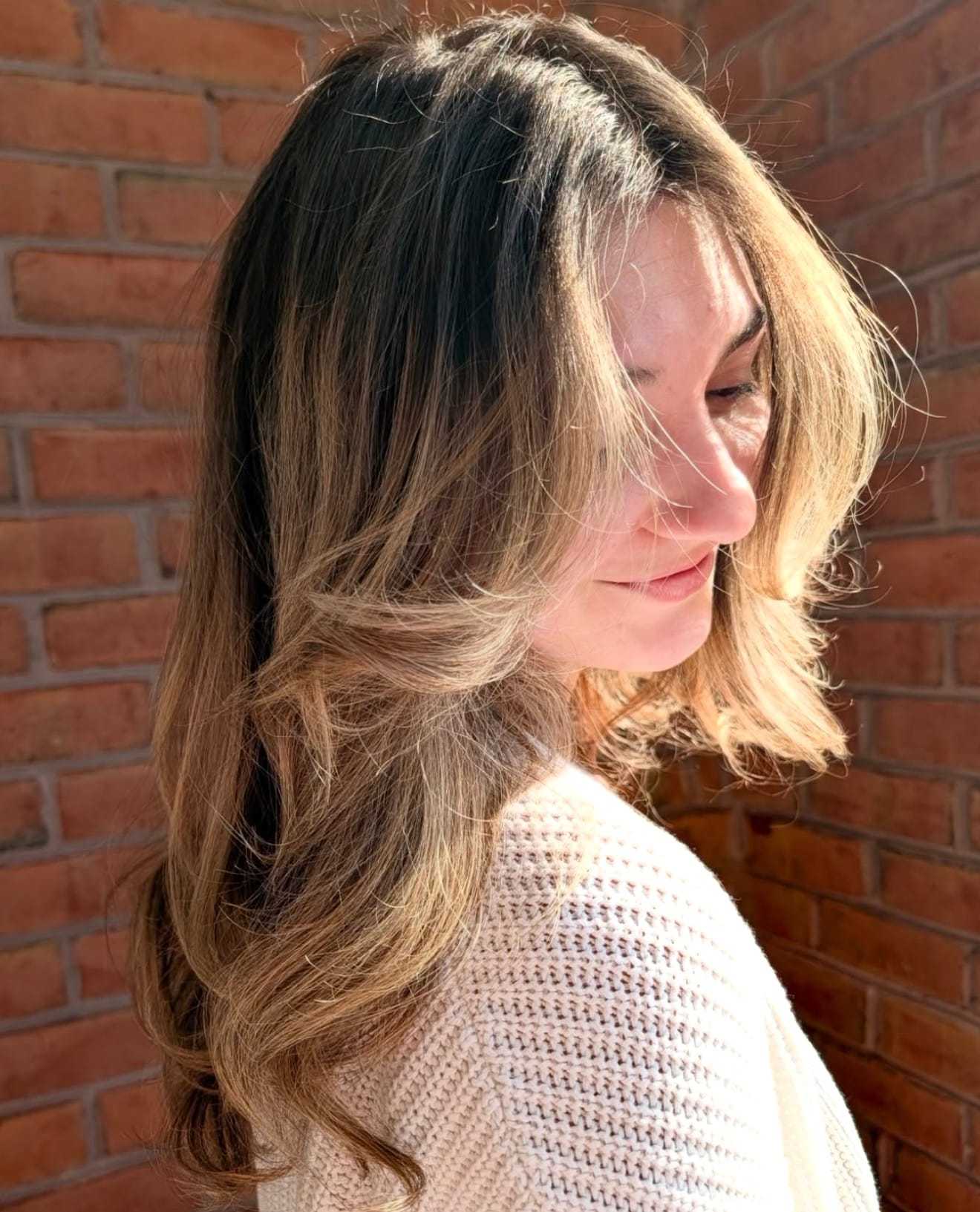 Woman with long, wavy hair stands against a brick wall, partially in sunlight.