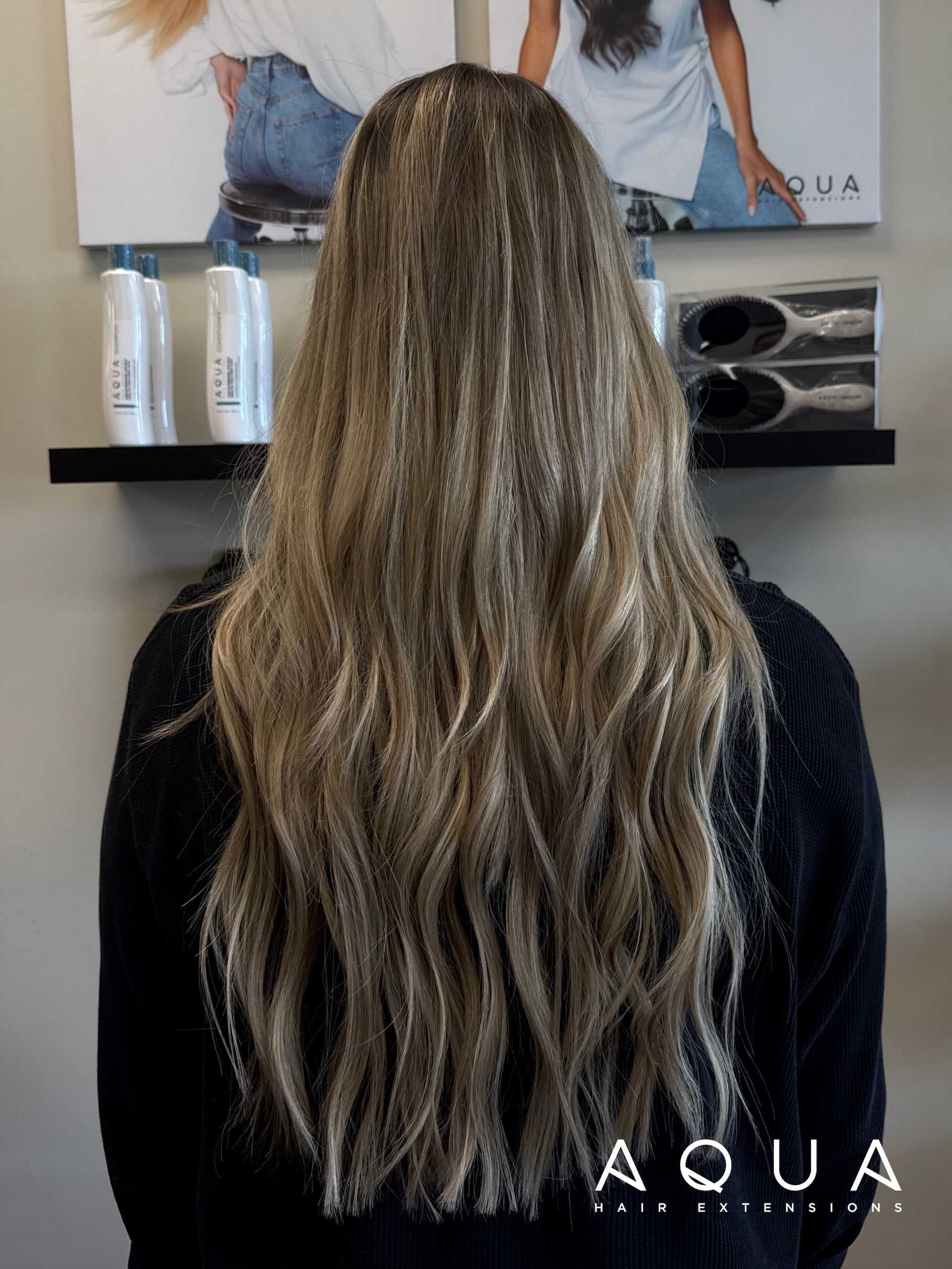 Long blonde hair with soft waves, displayed from the back. Hair products are visible on a shelf.