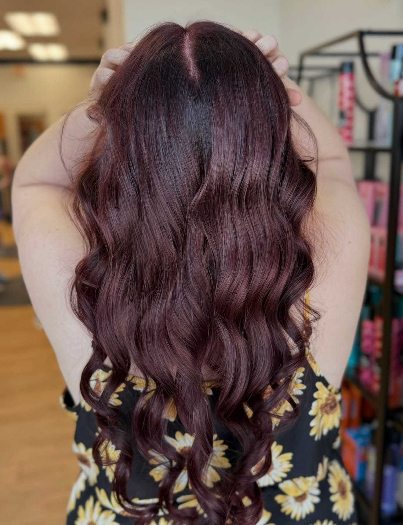 Back view of a person with long wavy burgundy hair, wearing a floral dress in a salon.