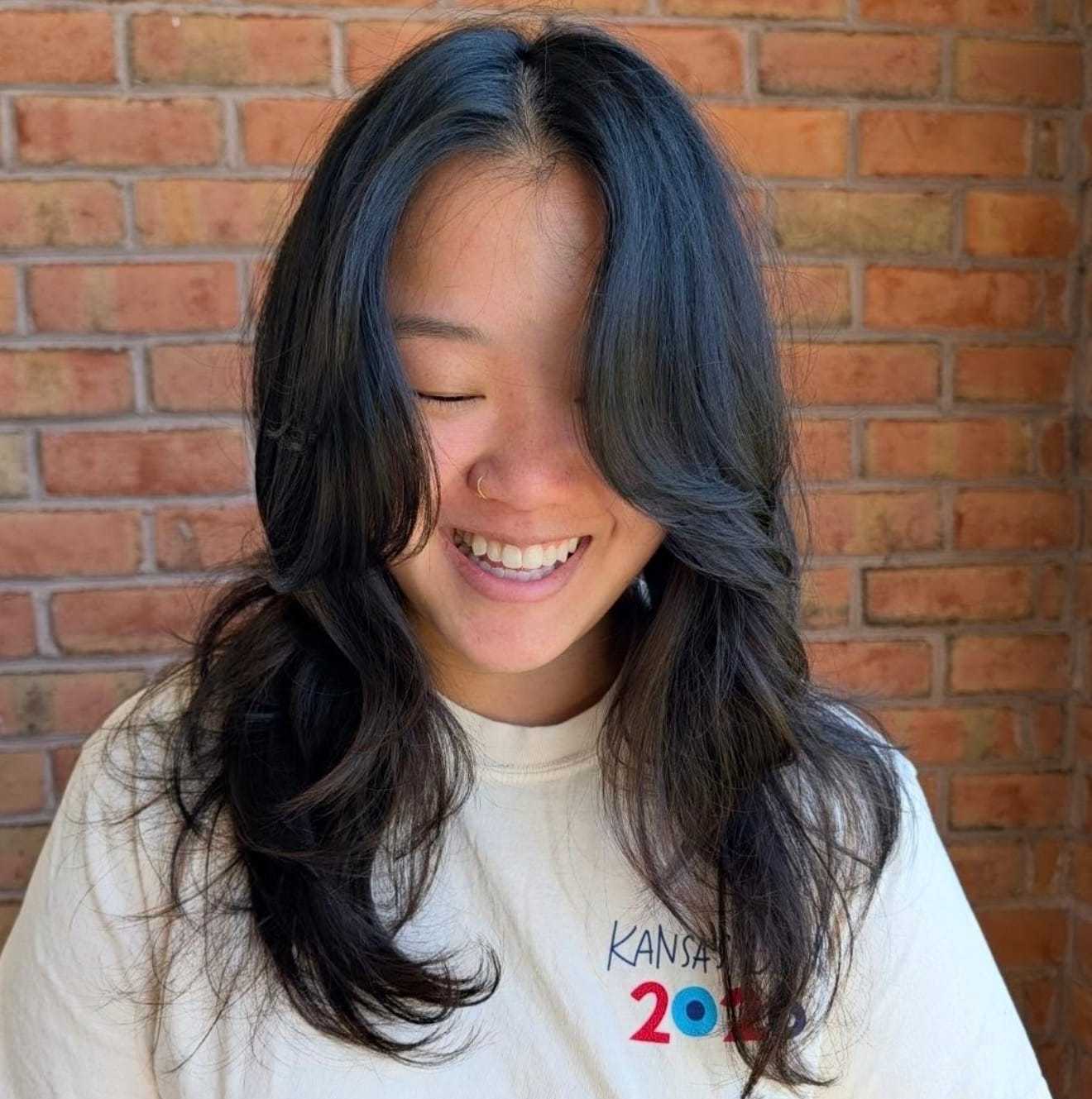 Asian woman with long, wavy hair smiling against a brick wall.