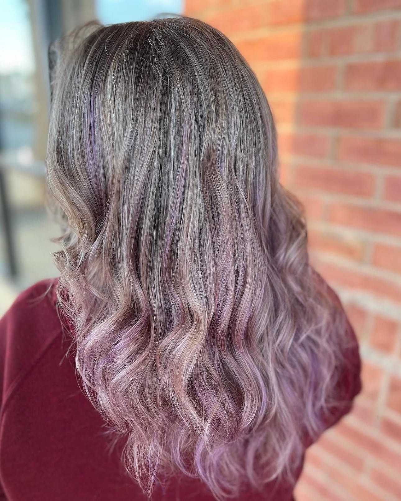 Wavy hair with lavender highlights cascading down to shoulders, against a brick wall backdrop.