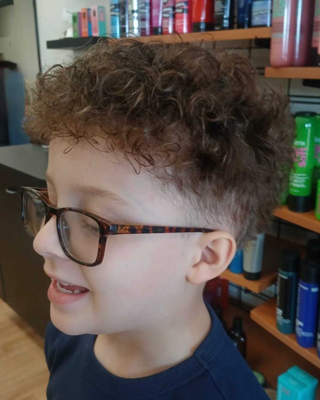 Young boy with curly hair and glasses in a salon, smiling.