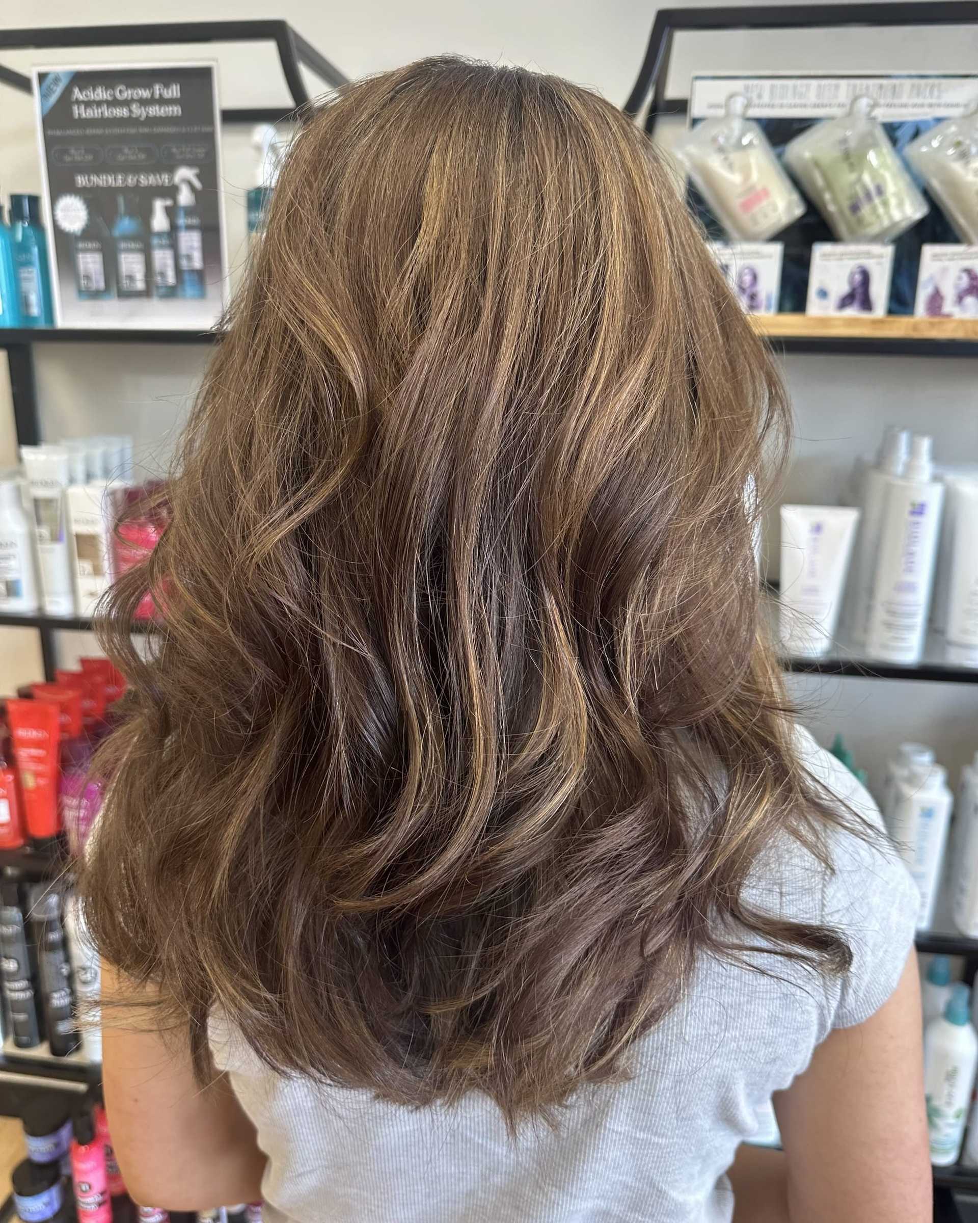 Brown-haired person with wavy layers stands in front of salon shelves with hair products.
