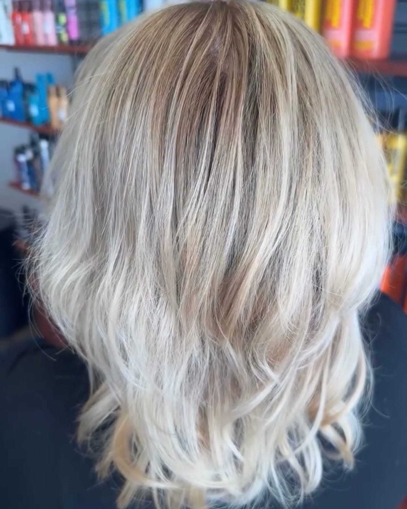 Blonde wavy hair from the back, salon shelves with colorful products in the background.