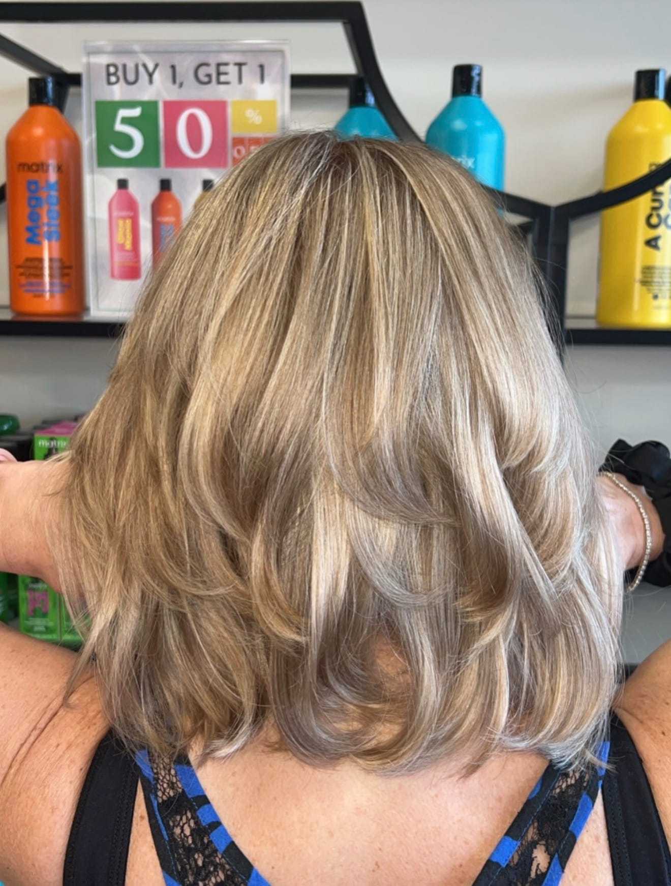 Back view of a woman with highlighted blonde hair in front of salon hair product display.