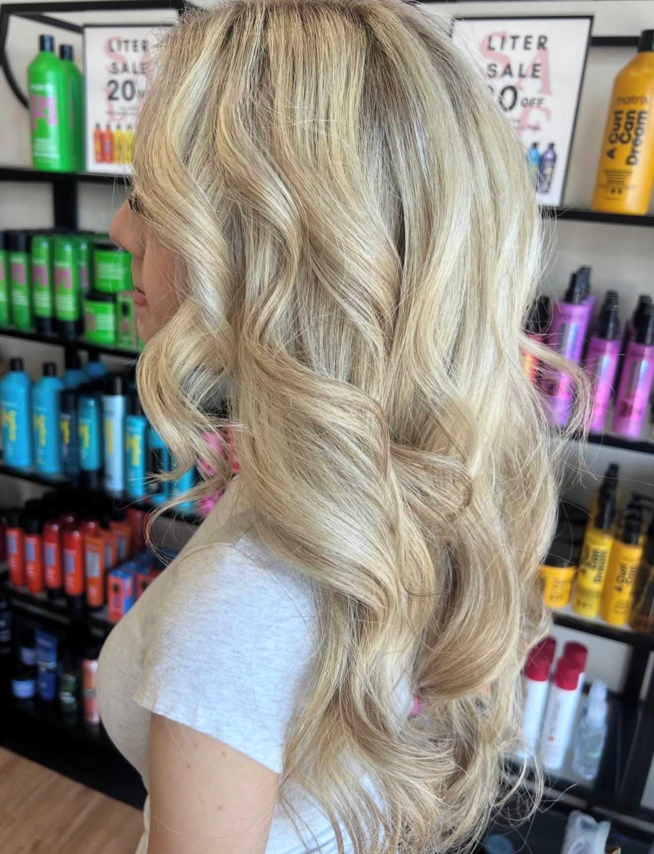 Blonde woman with wavy hair in a salon, colorful hair products displayed in the background.