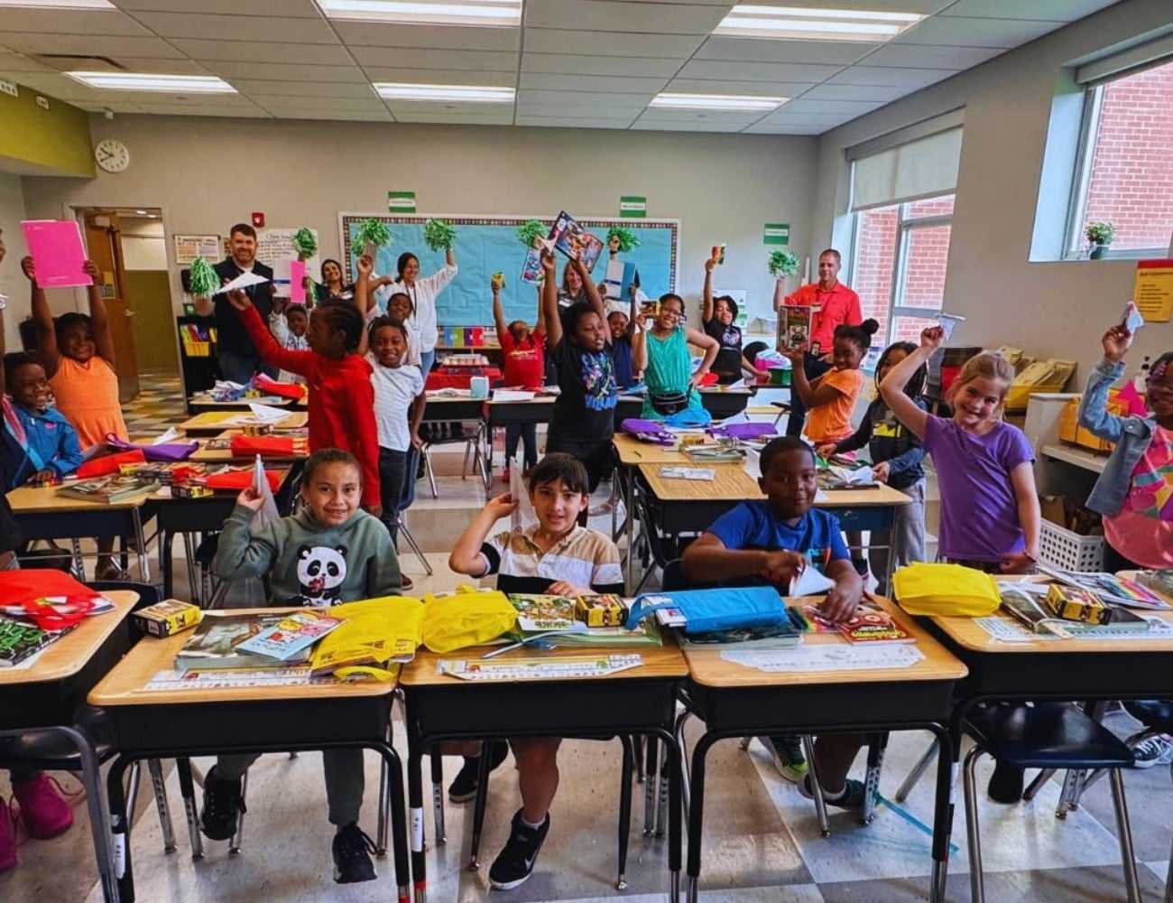 Students in a classroom holding up books, smiling, with teachers standing at the back.