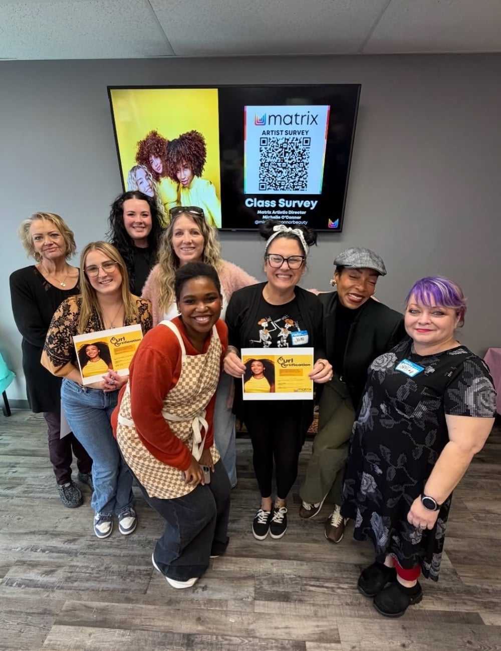 A group of smiling people posing indoors, holding flyers, with a survey QR code on a screen behind.