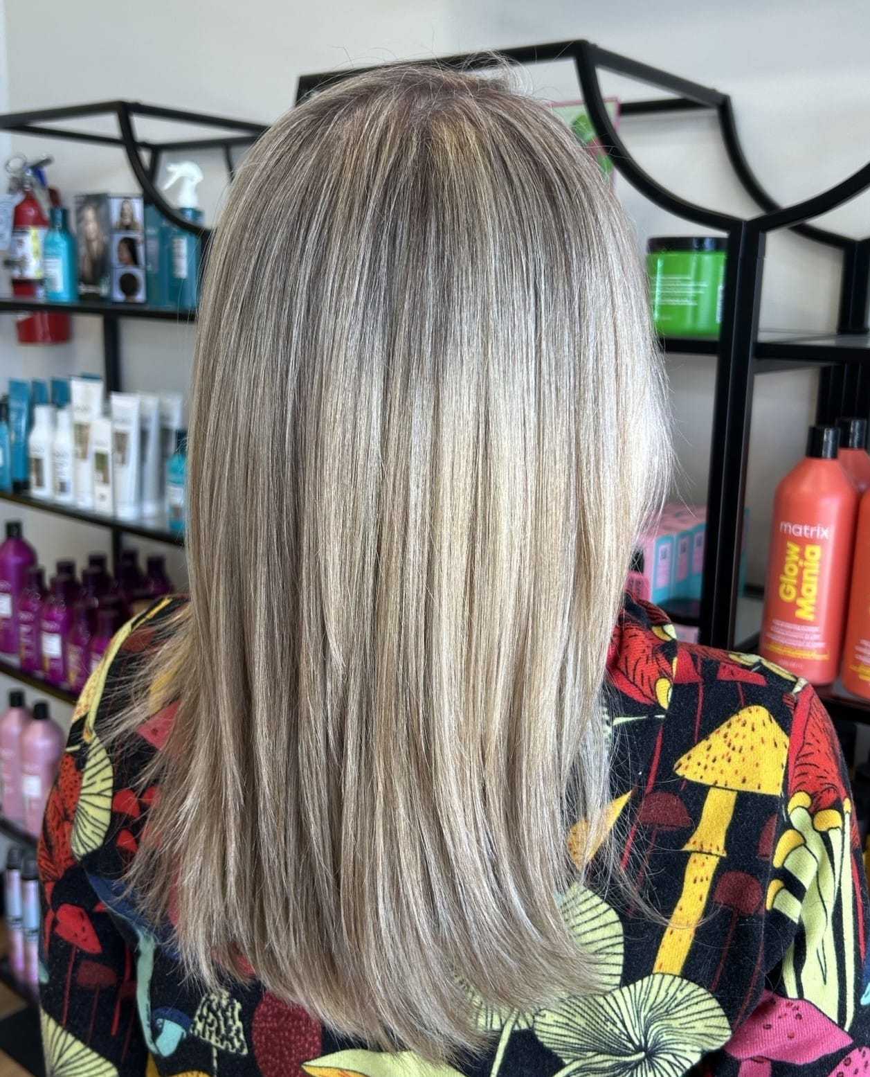 Blonde hair with highlights in a salon, styling products displayed on shelves behind.