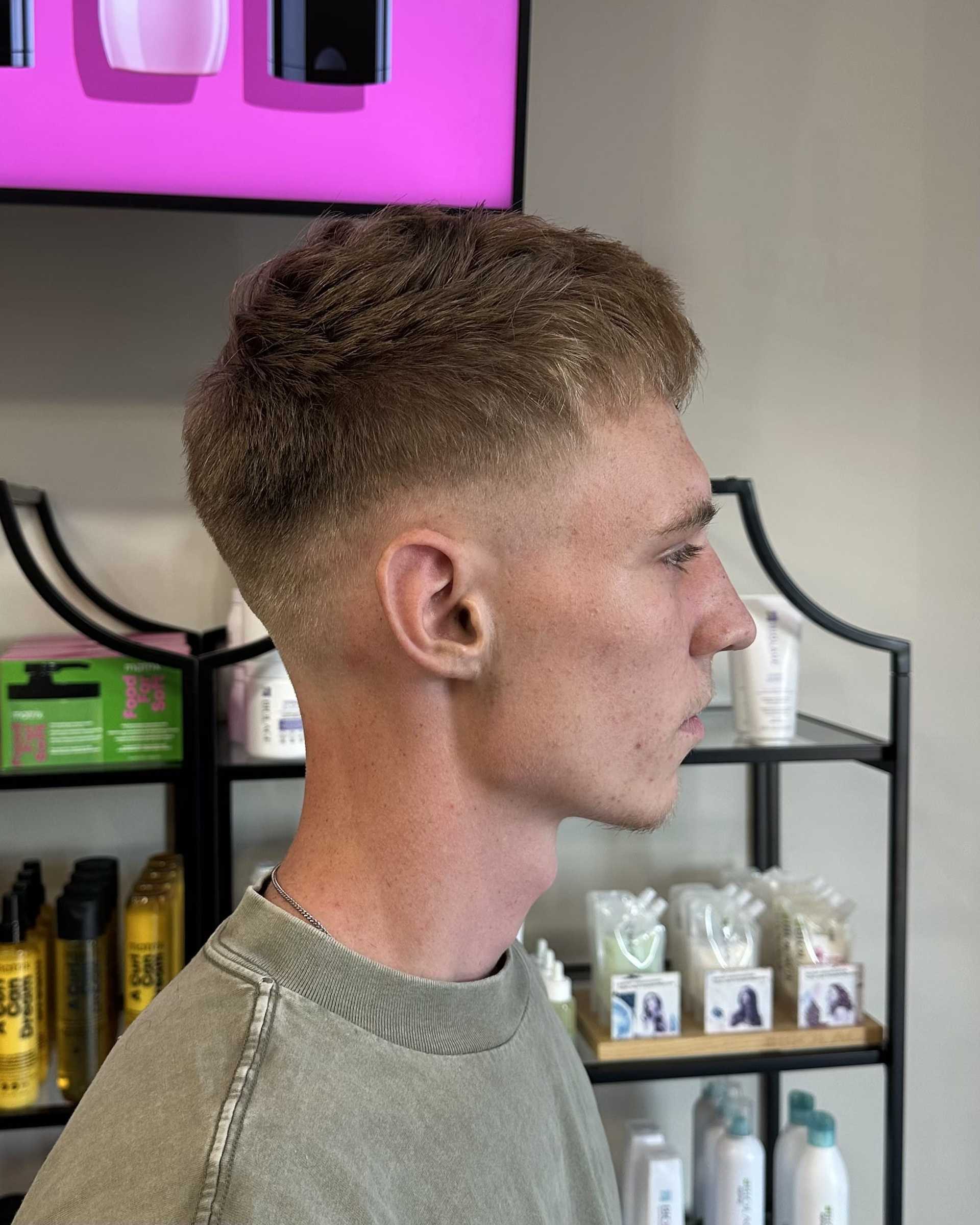 Young man with a short, tidy hairstyle in a salon, shelves of hair products visible behind him.